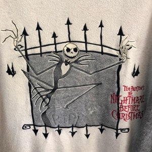 K&ouml;nnte beinhalten: Wei&szlig;er Fleece-Pullover mit einem schwarzen und grauen gestickten Bild von Jack Skellington aus Tim Burtons The Nightmare Before Christmas. Das Bild zeigt Jack Skellington, der auf einem Grabstein mit der Aufschrift "RIP" steht. Das Bild ist von einem schwarzen Zaun mit spitzen Oberteilen umgeben.