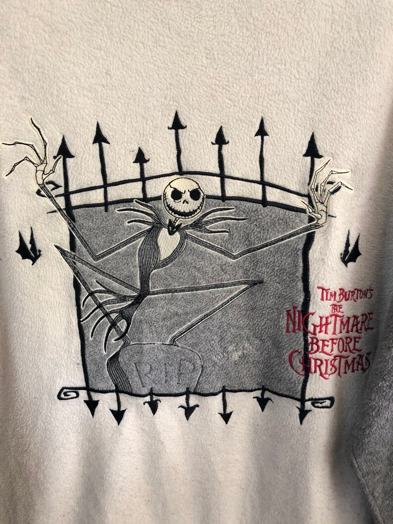 K&ouml;nnte beinhalten: Wei&szlig;er Fleece-Pullover mit einem schwarzen und grauen gestickten Bild von Jack Skellington aus Tim Burtons The Nightmare Before Christmas. Das Bild zeigt Jack Skellington, der auf einem Grabstein mit der Aufschrift "RIP" steht. Das Bild ist von einem schwarzen Zaun mit spitzen Oberteilen umgeben.