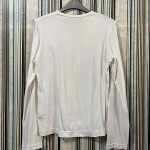 May include: A white long-sleeve shirt hanging on a gray hanger. The shirt is made of a soft material and has a crew neck. The sleeves are long and end at the wrists. The shirt is plain and has no visible design.