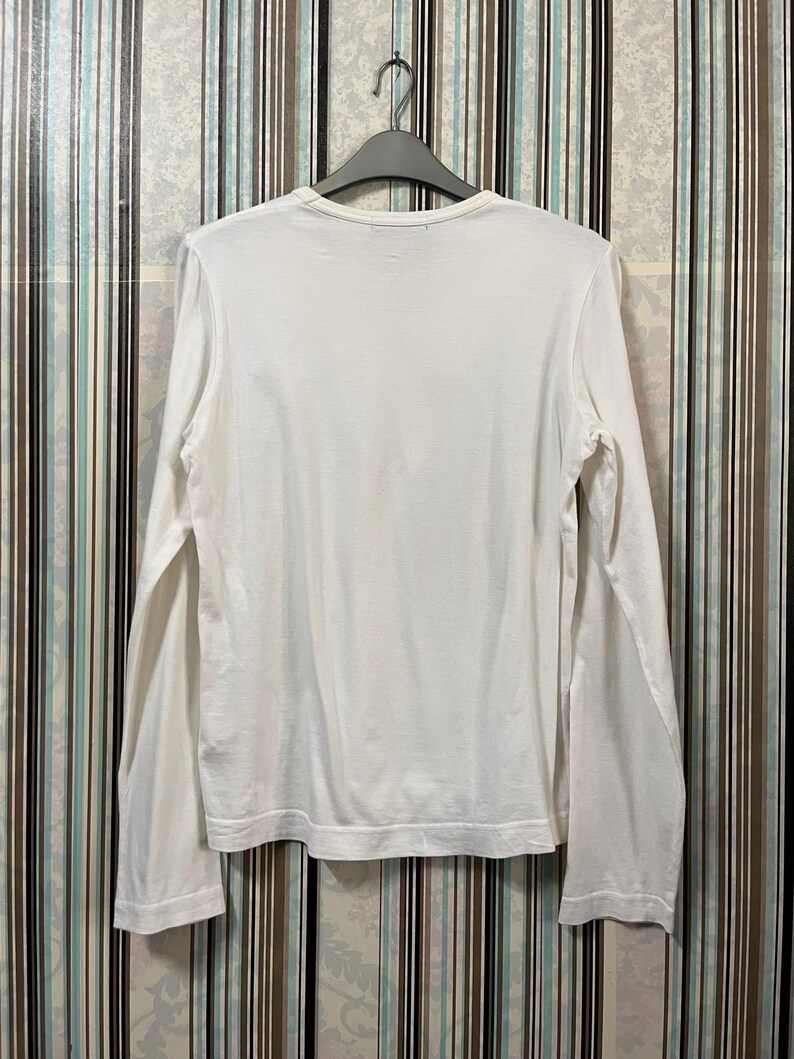 May include: A white long-sleeve shirt hanging on a gray hanger. The shirt is made of a soft material and has a crew neck. The sleeves are long and end at the wrists. The shirt is plain and has no visible design.