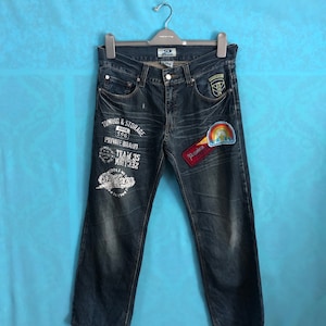 Peut inclure: Un jean en denim bleu foncé avec plusieurs patchs et des motifs imprimés. Les patchs comprennent un arc-en-ciel, un patch rouge et blanc avec le mot "Premium" et un patch vert avec une insigne militaire. Les motifs imprimés comprennent "Towing & Storage", "Dock 596", "Private Brand", "Team 35" et "Middle Air Town".