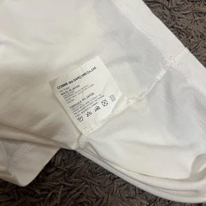 May include: Close-up of a white garment with a tag from COMME des GAR&Ccedil;ONS Co., Ltd. The tag indicates the item is made in Japan from 100% cotton. Washing instructions are also visible on the tag.
