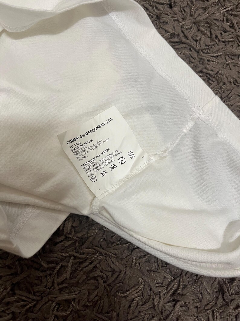 May include: Close-up of a white garment with a tag from COMME des GAR&Ccedil;ONS Co., Ltd. The tag indicates the item is made in Japan from 100% cotton. Washing instructions are also visible on the tag.