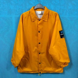 Puede incluir: Una chaqueta de entrenador de nylon amarilla con cierre de presión negro y un parche de logotipo azul y negro que dice "Helly Hansen" en la manga izquierda.