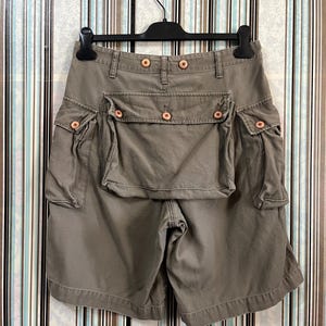 May include: Olive green cargo shorts with multiple pockets and wooden button closures. The shorts are hanging on a black hanger against a striped background. The shorts have a high waist and a relaxed fit.
