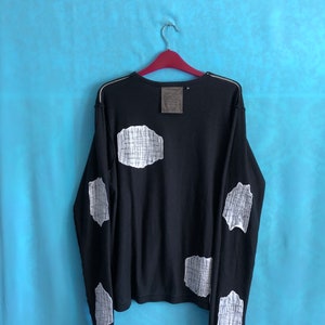 Può includere: Maglietta nera a maniche lunghe con toppe a griglia bianca sulle maniche e sulla schiena. La maglietta ha un collo rotondo e un'etichetta sul retro.