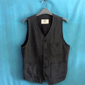 May include: A dark grey waistcoat with a button fastening and two large pockets on the front. The waistcoat is made from a textured fabric and has a label that reads "The Workers Club" and "Made in China".