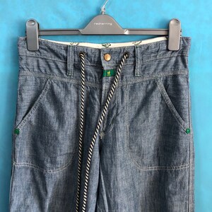 May include: Blue denim trousers with a white and black drawstring and green accents. The trousers have two front pockets and two back pockets.