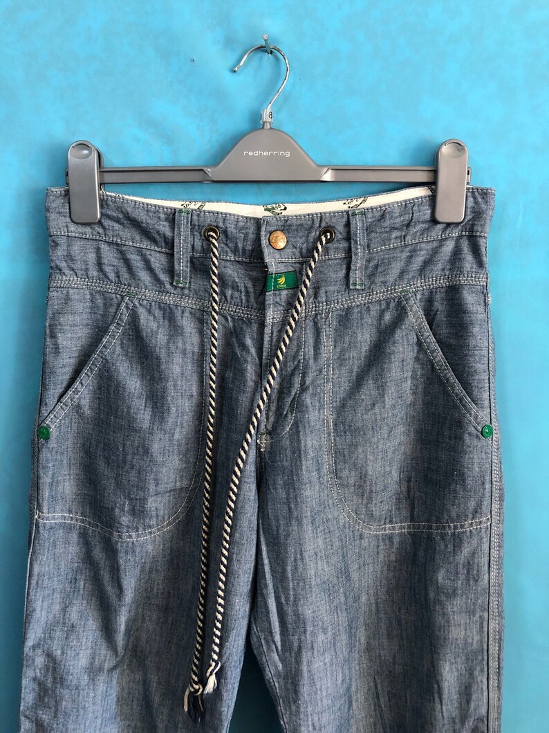 May include: Blue denim trousers with a white and black drawstring and green accents. The trousers have two front pockets and two back pockets.
