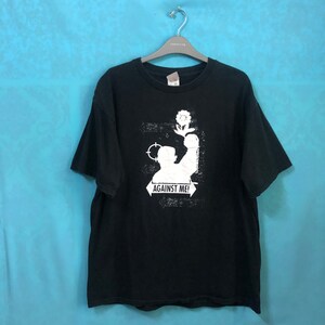 May include: Black t-shirt with a white graphic featuring a figure holding a flower and the text "AGAINST ME!". The shirt is hanging on a metal hanger against a blue background.