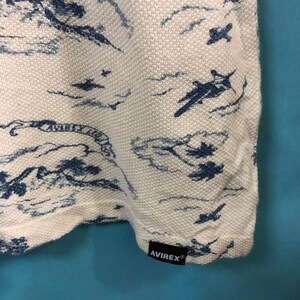 May include: White t-shirt with a blue and white print of airplanes flying over clouds. The shirt has a black tag with the word "AVIREX" on it.