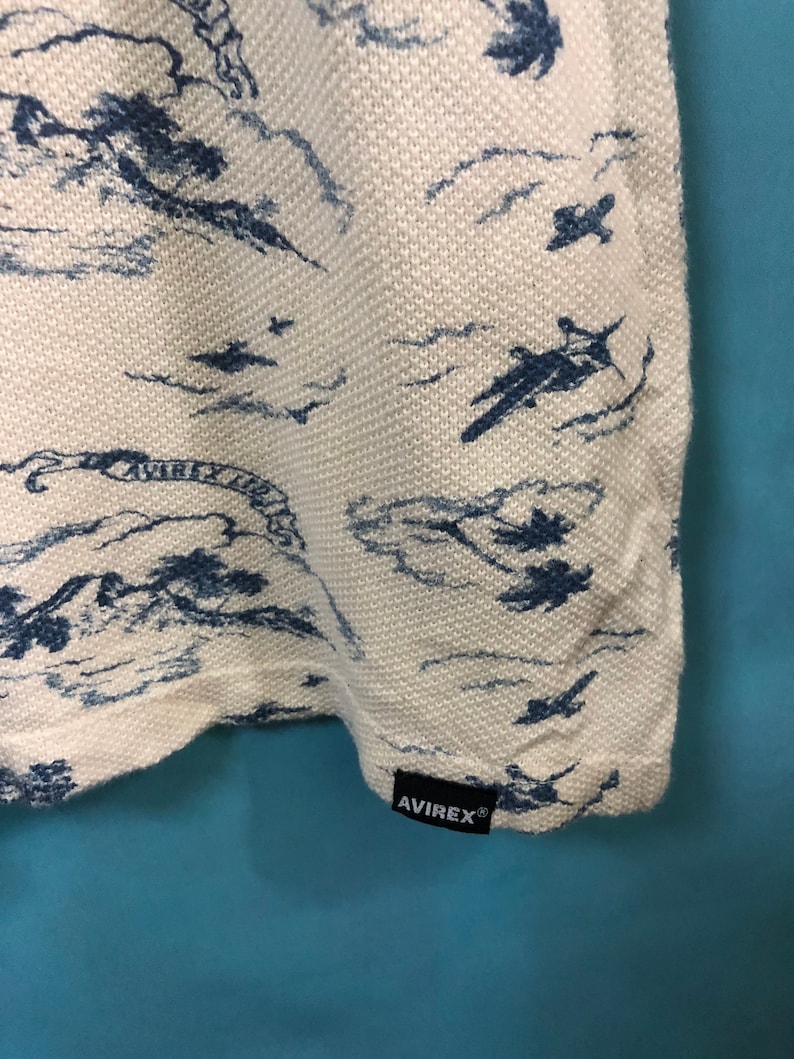 May include: White t-shirt with a blue and white print of airplanes flying over clouds. The shirt has a black tag with the word "AVIREX" on it.