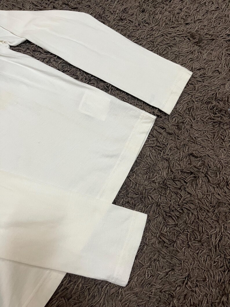 May include: A white long-sleeved shirt with a simple design. The shirt is made of a soft, likely cotton material. The sleeves and body of the shirt are visible, with a focus on the clean, minimalist aesthetic.