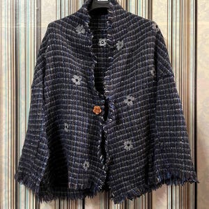 Pode incluir: Casaco de tweed azul marinho com bainha com franjas e enfeites florais. O casaco tem um fecho de botão de madeira e está pendurado num cabide preto.