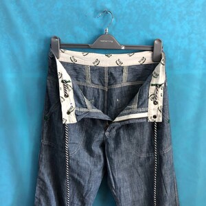 May include: A pair of blue denim jeans with a white and black rope detail hanging from the waist. The jeans have a white waistband with a repeated pattern of a green and white logo.