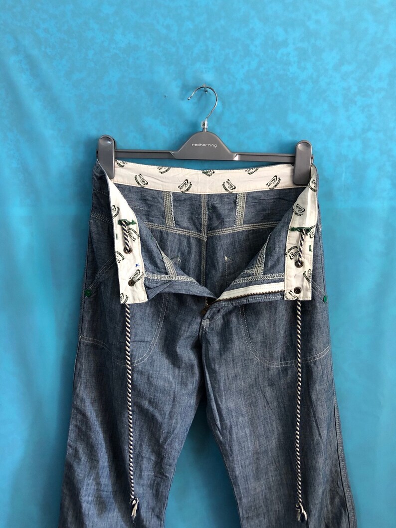 May include: A pair of blue denim jeans with a white and black rope detail hanging from the waist. The jeans have a white waistband with a repeated pattern of a green and white logo.