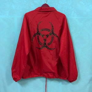 Puede incluir: Chaqueta cortavientos roja con un símbolo de biohazard negro en la espalda.
