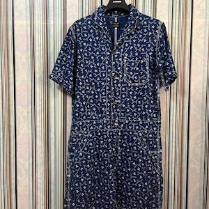 May include: A dark blue jumpsuit with a white floral pattern. The short-sleeved jumpsuit has a collared neckline, button-up front, and pockets. The garment is hanging on a black hanger.