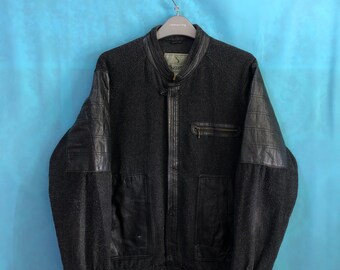 Puede incluir: Una chaqueta negra de cuero y lana con cierre de cremallera y cuello alto. La chaqueta tiene un parche de logotipo en el pecho izquierdo que dice "Bugatti".