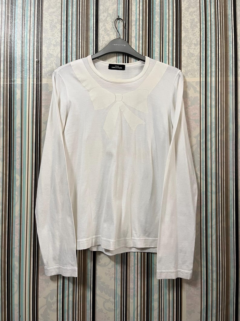 May include: White long-sleeve shirt with a large bow detail on the chest. The shirt is hanging on a black hanger. The shirt has a crew neck and is made of a soft material. The shirt is a solid white color.