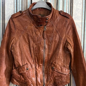 May include: A brown leather jacket with a stand-up collar, zipper closure, and two front pockets. The jacket has a textured appearance and is hanging on a hanger. The jacket's color is a warm, reddish-brown.
