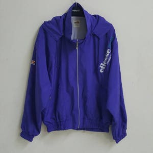 May include: A purple hooded jacket with a zipper closure and the word "ellesse" printed on the right side. The jacket has a white drawstring at the bottom and a small logo on the left sleeve.