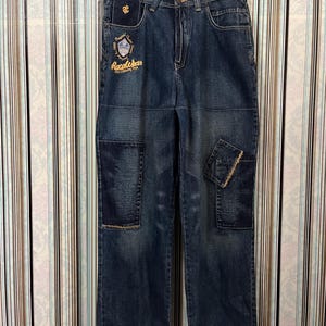Pode incluir: Jeans de ganga de lavagem escura com várias aplicações e um corte descontraído. Os jeans apresentam uma aplicação decorativa no bolso da frente e uma aplicação com o logótipo acima do bolso da frente. Os jeans estão pendurados num cabide de madeira.