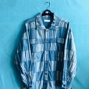 May include: Blue denim shirt with a patchwork design of vertical white stripes and solid blue sections. The shirt has a collared neckline, button-down front, and long sleeves. It hangs on a black hanger against a light blue background.