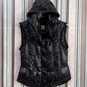 May include: Black sleeveless puffer vest with a zip closure and a hood. The vest has a shiny, quilted fabric and multiple pockets with buckles.