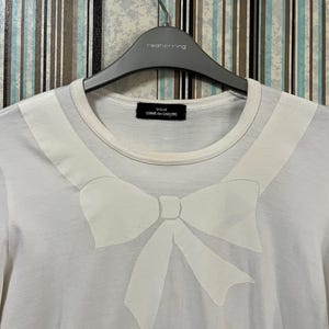 May include: White t-shirt featuring a large, light-colored bow design on the front. The shirt has a round neckline and short sleeves. The tag reads "tricot COMME des GAR&Ccedil;ONS". The shirt is hanging on a gray hanger.