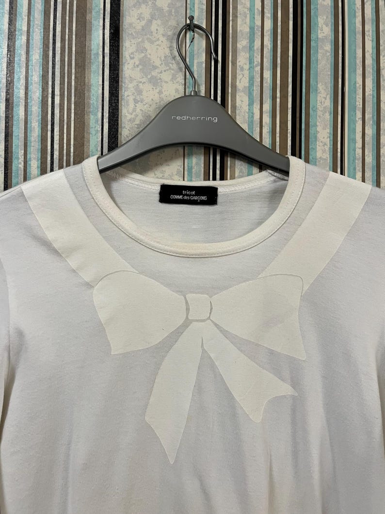 May include: White t-shirt featuring a large, light-colored bow design on the front. The shirt has a round neckline and short sleeves. The tag reads "tricot COMME des GAR&Ccedil;ONS". The shirt is hanging on a gray hanger.