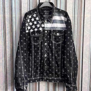 May include: A black denim jacket with a white star pattern and an American flag design on the front. The jacket has a button closure and two pockets.