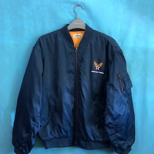 May include: A navy blue bomber jacket with a zipper closure and a patch on the left chest that reads "Army Air Forces" with a star and wings design.