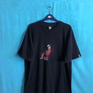 Puede incluir: Camiseta negra con un estampado gráfico de una persona con un atuendo rojo y negro sentada en una silla. El gráfico está rodeado por un patrón geométrico.