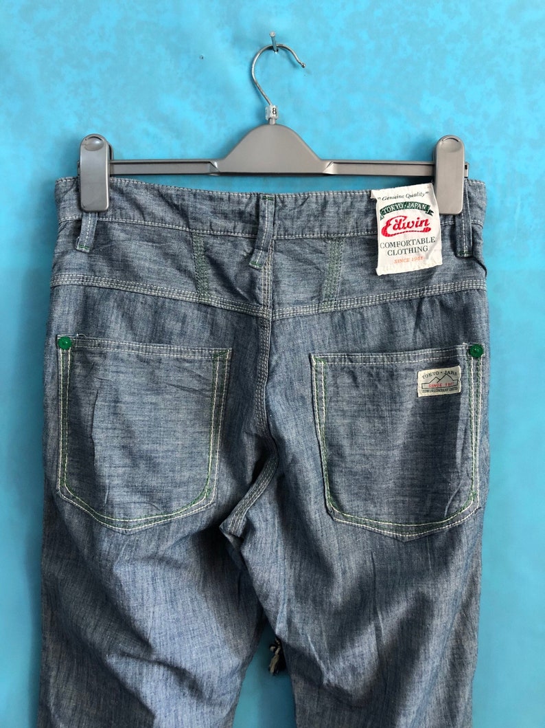 May include: Blue denim jeans with green stitching and a white label that says "Edwin Comfortable Clothing Since 1961" hanging on a black hanger.