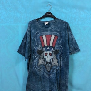 Könnte beinhalten: Ein blaues Batik-T-Shirt mit einem Motiv eines Schädels, der einen rot-weiß-blauen Onkel-Sam-Hut trägt. Der Schädel ist von einem kreisförmigen Design umgeben.