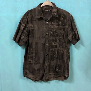 May include: A black short-sleeved button-down shirt with a striped pattern and a print of text and symbols. The shirt has a button-up collar and a pocket on the left side. The text on the shirt includes "Only", "Anarchists", "Are", "SANG", "ROUS", "1600", "SONY", and "A".