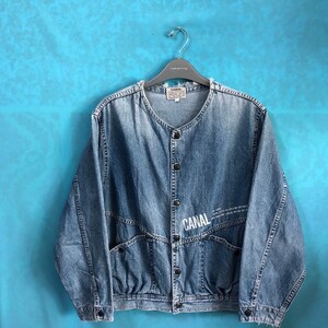 May include: A light blue denim jacket with a button closure and a white "CANAL" logo on the front. The jacket has a relaxed fit and two large pockets on the front.