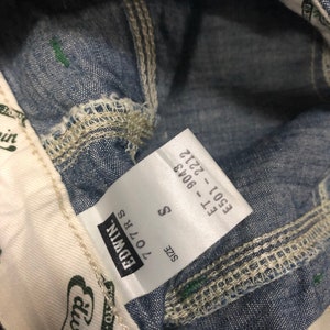 May include: A white tag with black text that reads "SIZE S 170/78 EDWIN" and "E501 - F11 2722 - 8043". The tag is attached to a pair of blue denim jeans.
