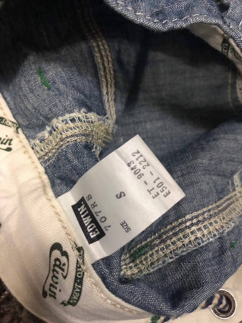May include: A white tag with black text that reads "SIZE S 170/78 EDWIN" and "E501 - F11 2722 - 8043". The tag is attached to a pair of blue denim jeans.