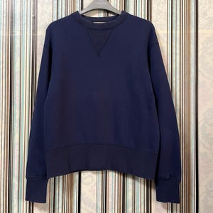 Peut inclure: Un sweat-shirt bleu marine avec un col ras du cou et des manches longues. Le sweat-shirt présente un détail en forme de V à l'encolure et des poignets et un ourlet côtelés. Le vêtement est suspendu à un cintre.