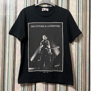 Może przedstawiać: Czarny t-shirt z czarno-białym zdjęciem osoby na scenie i napisem "THE FUTURE IS UNWRITTEN" w białym prostokącie. Koszulka wisi na wieszaku.
