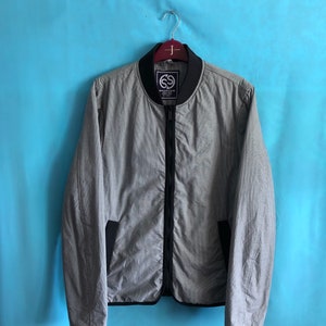 May include: A gray bomber jacket with a black zipper and trim, hanging on a wooden hanger. The jacket has a subtle pattern and black pockets. The label on the collar reads "SPORTICA".