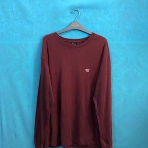 Könnte beinhalten: Ein langärmeliges burgunderfarbenes T-Shirt mit einem kleinen roten Herz-Motiv auf der linken Brust.