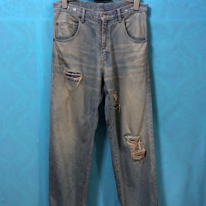 Puede incluir: Un par de pantalones vaqueros de mezclilla azul claro con detalles desgastados, que incluyen varios agujeros y bordes deshilachados. Los pantalones vaqueros están colgados de una percha negra sobre un fondo azul.