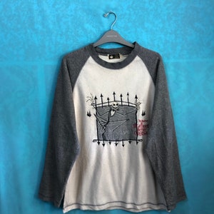 K&ouml;nnte beinhalten: Ein wei&szlig;es und graues Langarmshirt mit Baseball-Design. Das Shirt zeigt eine Grafik von Jack Skellington aus dem Film "Nightmare Before Christmas" mit dem Filmtitel unter der Grafik.