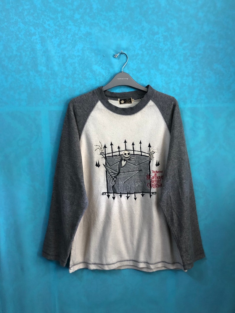 K&ouml;nnte beinhalten: Ein wei&szlig;es und graues Langarmshirt mit Baseball-Design. Das Shirt zeigt eine Grafik von Jack Skellington aus dem Film "Nightmare Before Christmas" mit dem Filmtitel unter der Grafik.
