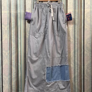 May include: A light gray maxi skirt with a drawstring waist and a denim patch. The skirt is hanging on a wooden hanger against a striped background. The skirt has pockets and a relaxed fit.