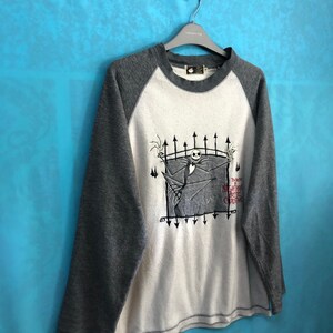 K&ouml;nnte beinhalten: Ein wei&szlig;es und graues Langarmshirt mit Baseball-Design. Das Shirt zeigt eine Grafik von Jack Skellington aus dem Film "Nightmare Before Christmas" mit dem Text "The Nightmare Before Christmas".