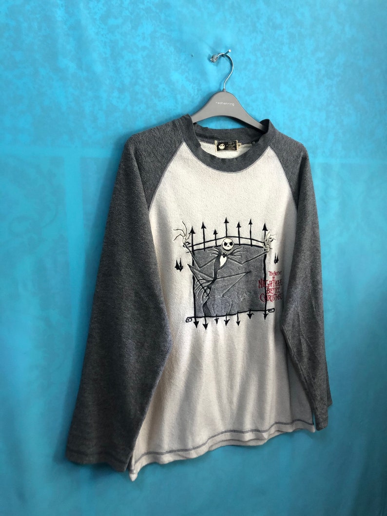 K&ouml;nnte beinhalten: Ein wei&szlig;es und graues Langarmshirt mit Baseball-Design. Das Shirt zeigt eine Grafik von Jack Skellington aus dem Film "Nightmare Before Christmas" mit dem Text "The Nightmare Before Christmas".